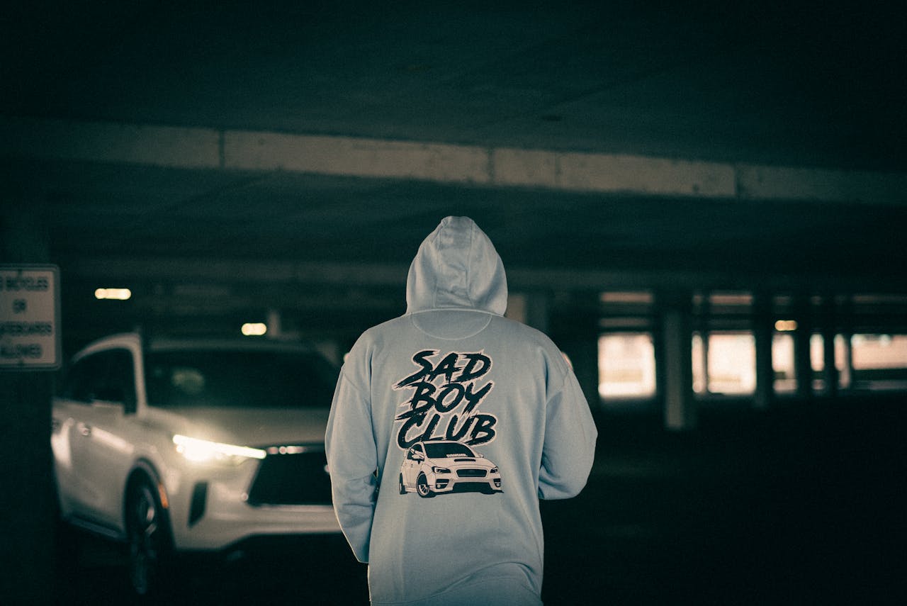 Person wearing a 'Sad Boy Club' hoodie walking near a car in an indoor parking garage.