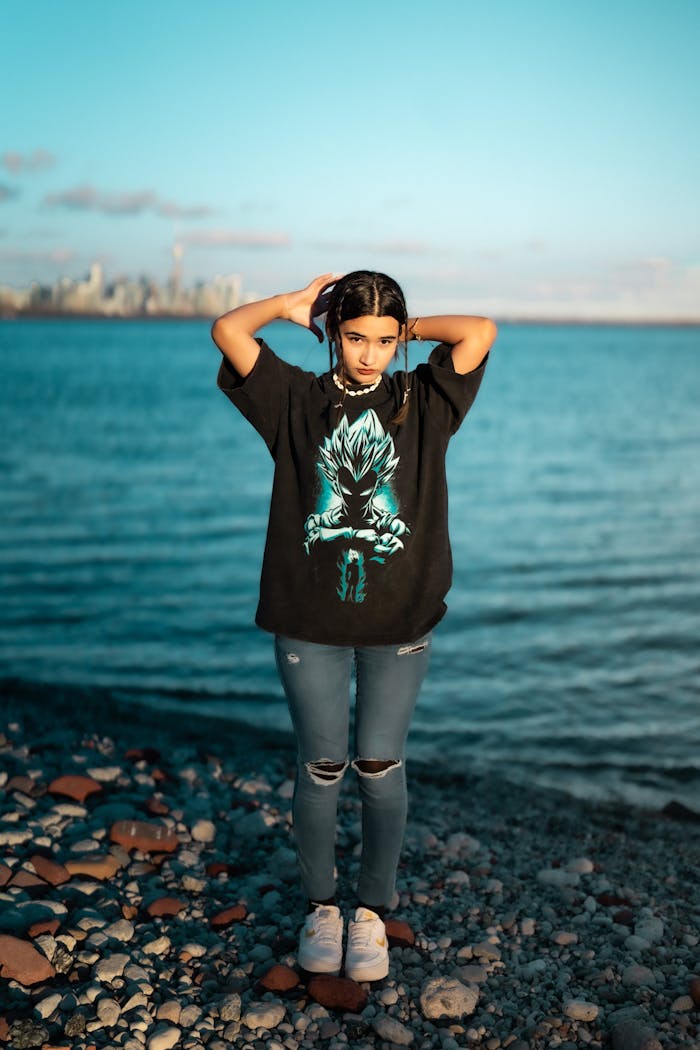 A teenager standing by the water with a city skyline in the background during sunset.