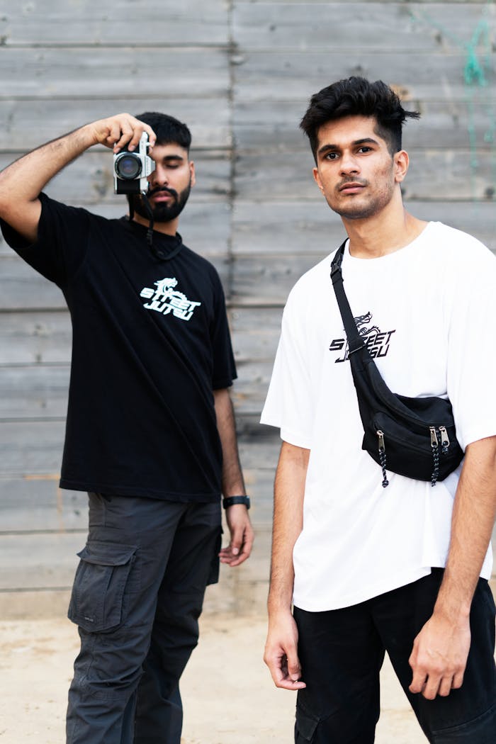 Two young men in casual wear, one holding a camera, posing against a rustic wooden backdrop.