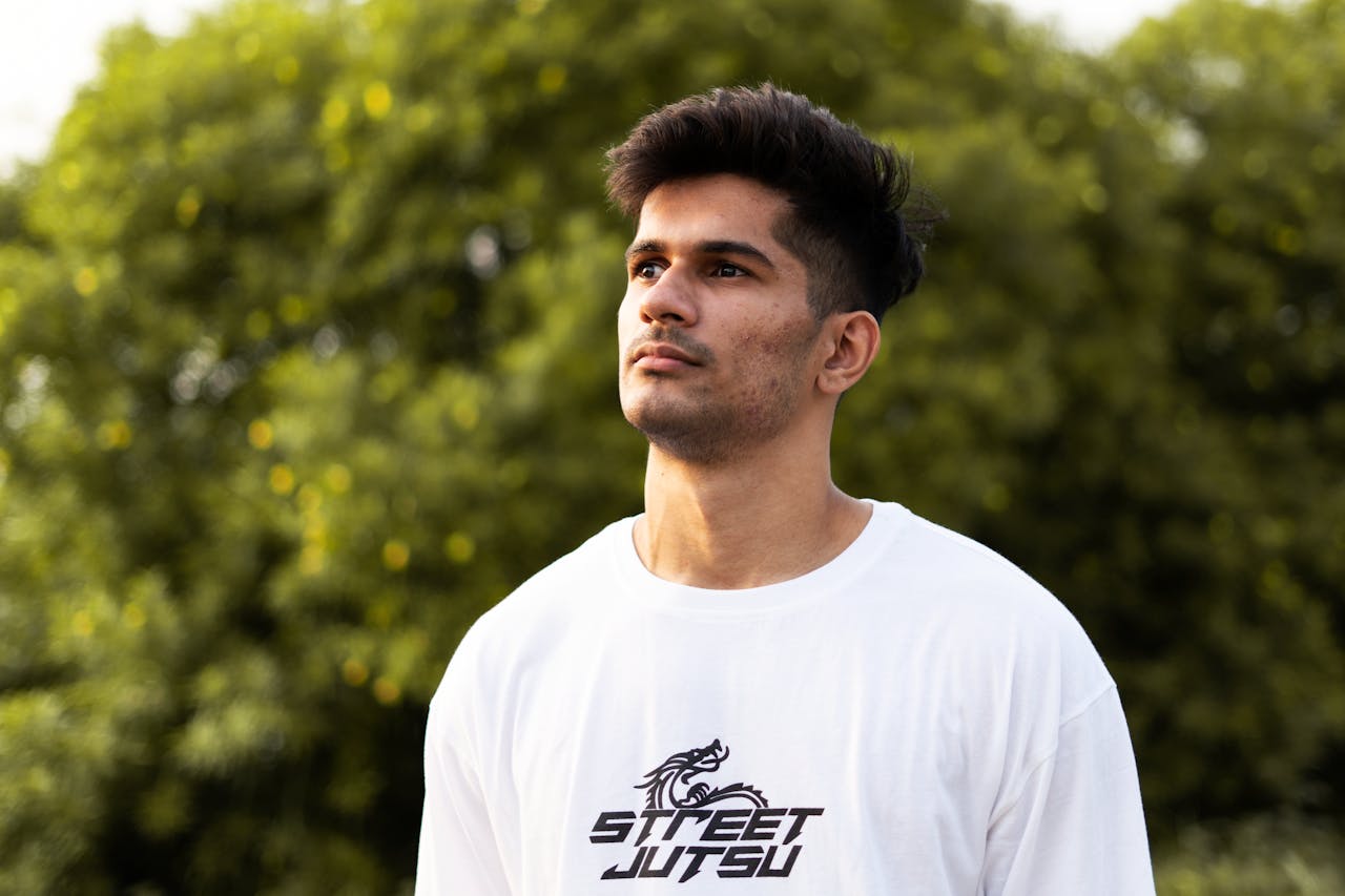 Young man in a white 'Street Jutsu' shirt looking upwards outdoors, with blurred green background.